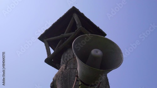 Vintage loudspeaker mounted on a rustic wooden pole broadcasting an announcement against a clear blue sky, symbolizing propaganda, public address, or emergency alerts from the past
