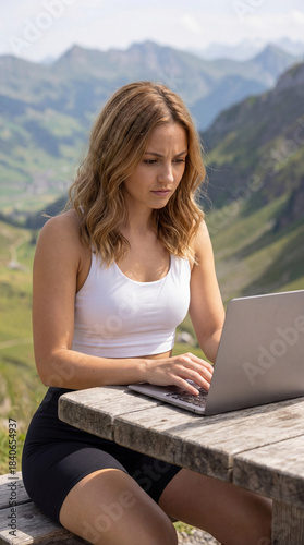 Concentrated young Swiss woman typing on her laptop on a sunny mountain terrace with a scenic alpine view in the background. Modern remote work and flexible workplace concept in nature.