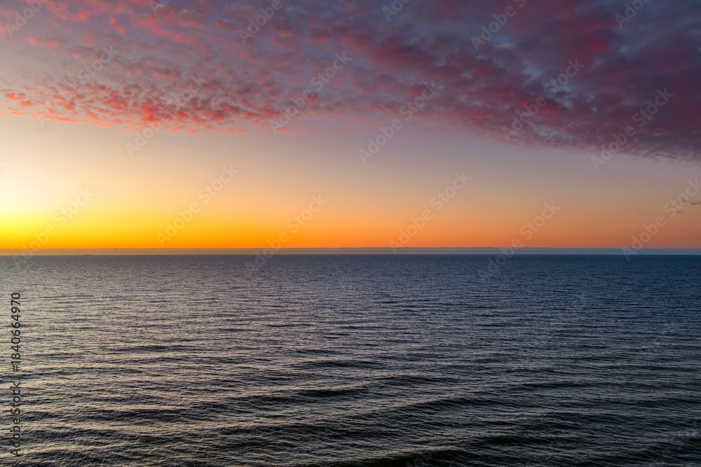 Fototapeta premium Aerial view of Baltic Sea coast in winter, with bare trees on the cliff and waves rolling under a vivid red sunset sky. Stark northern beauty, elemental in fading light.