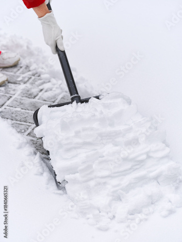 A person wearing white gloves using a black snow shovel to clear fresh white snow from a paved outdoor area. Shallow depth of field.