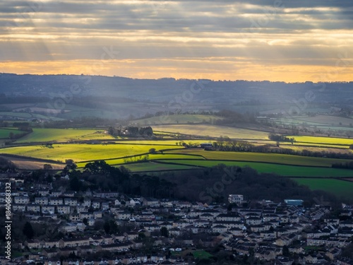 Aerial view of sunbeams pierce through a cloudy sky, illuminating the green fields beyond the city, creating a mesmerizing contrast with the dark cityscape., Bath, England, United Kingdom.