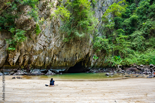 Person sitting on the sand of a hidden beach, observing the shimmering emerald water by the entrance of Tham Morakot cave, surrounded by lush tropical vegetation and towering limestone cliffs