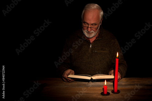 An older man with gray hair sits in the soft glow of candlelight, absorbed in a book. The dark background adds to the peaceful, intimate atmosphere, highlighting the calm of the quiet moment.