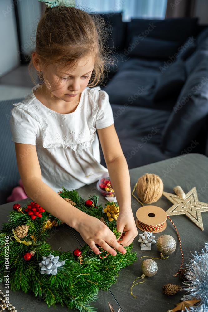 Naklejka premium Little girl finishing her handmade Christmas wreath by tying a string to hang it. Final step of a festive DIY project captured at home in a cozy family setting.