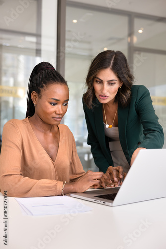 Young Latin mentor training new African employee at laptop, pointing at screen, explaining application work, giving feedback, advice, professional support, consultation. Vertical shot