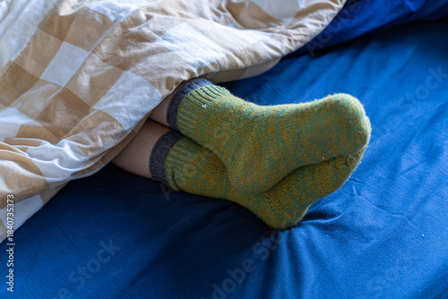 Person resting under a blanket with warm wool socks sticking out on a blue bed, cozy winter morning atmosphere.