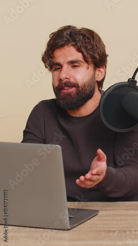 Happy blogger man recording podcast at table, giving workshop, webinar. Young Indian male speaking at microphone and laptop webcam, making video call. Handsome Hindu guy isolated on beige background.