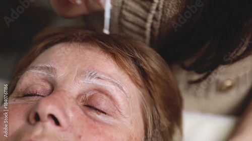 A young cosmetologist colors the eyebrows of a senior woman.