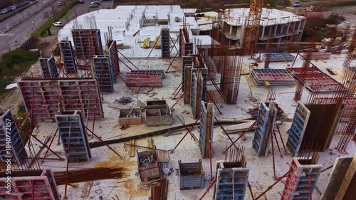 Heavy machinery and workers fill the construction site as they skillfully erect a new building. Scaffolding and rebar create a vivid scene under the bright afternoon sun.