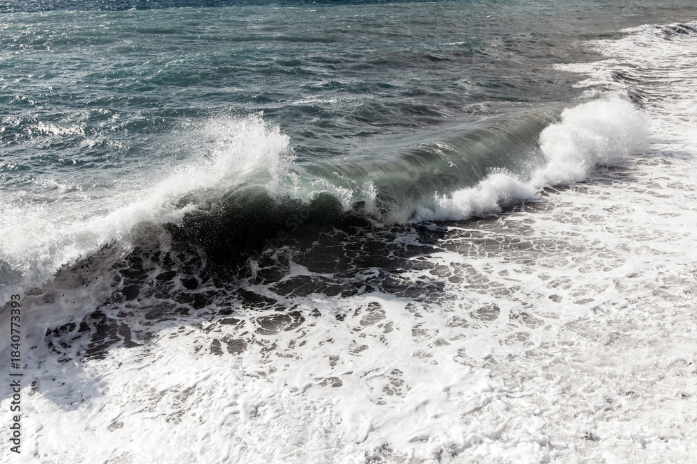 Fototapeta premium Powerful ocean wave crashing onto sandy shore, creating frothy white foam, with glistening water reflecting sunlight, embodying the beauty of nature's force and tranquility