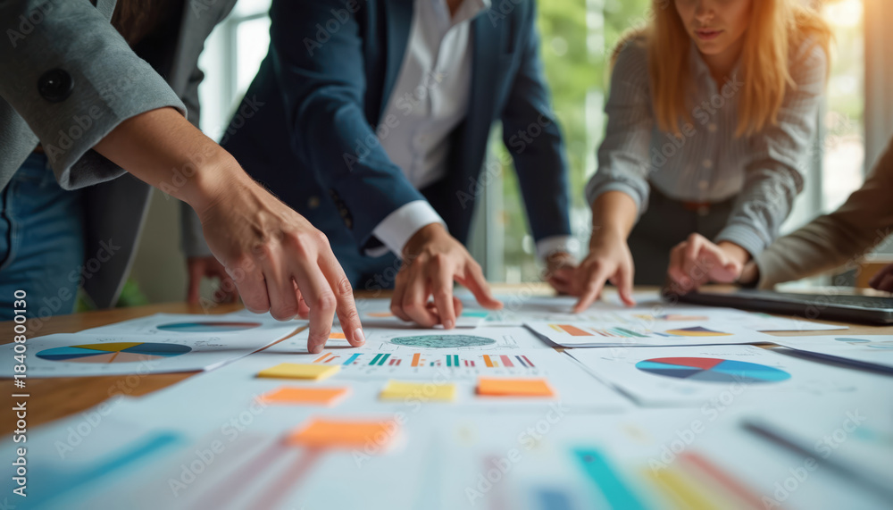Fototapeta premium Team collaborates discussing charts and graphs on desk. People point at papers, analyzing data for project success. Colleagues share ideas in modern office setting, planning future strategy.