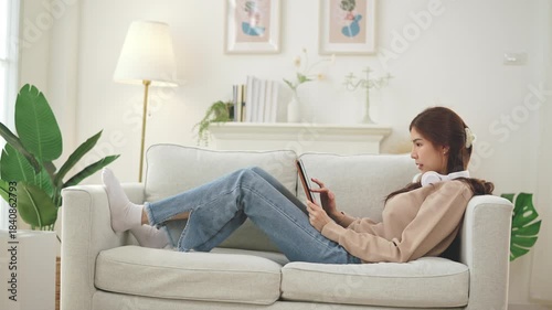Young Asian Woman Relaxing on Sofa Using Digital Tablet in modern living room at Home. She is wearing casual attire and has headphones around her neck. home comfort, digital lifestyle.
