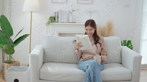 Relaxed young Asian woman reading magazine or book and drinking coffee on sofa at home. the scene emphasizes relaxation, comfort, hygge, and a peaceful indoor lifestyle.