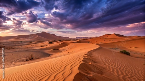 Fototapeta Naklejka Na Ścianę i Meble -  Dramatic sunset over vast orange sand dunes in the desert landscape with intense purple and dark clouds in the sky