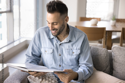 Принти на полотні Cheerful young Hispanic man sits comfortably on couch reading large book and smiling with joy, looks relaxed and contented, enjoying quiet and peaceful moment in cozy living room