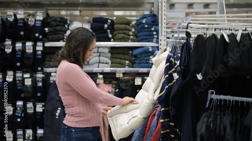 Woman choosing white winter vest in supermarket clothing section, holding the padded sleeveless jacket and checking fit and warmth, shopping and seasonal fashion concept