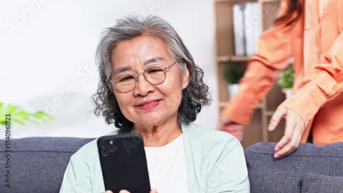 Asian Elderly woman and adult daughter smiling while video calling on smartphone in cozy living room with glasses on sofa bookshelf plant warm family moment affectionate interaction joyful expression