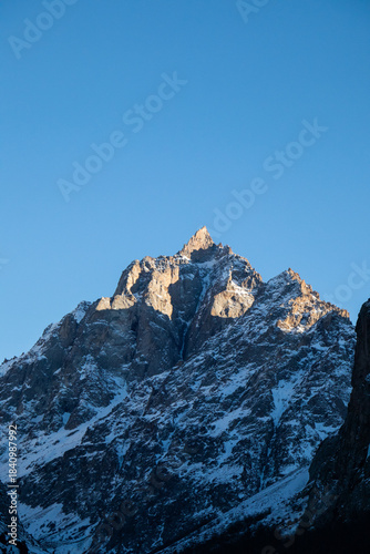 Extreme close-up of a rugged, rocky mountain peak dusted with snow, dramatically lit by low winter sun against a clear blue sky