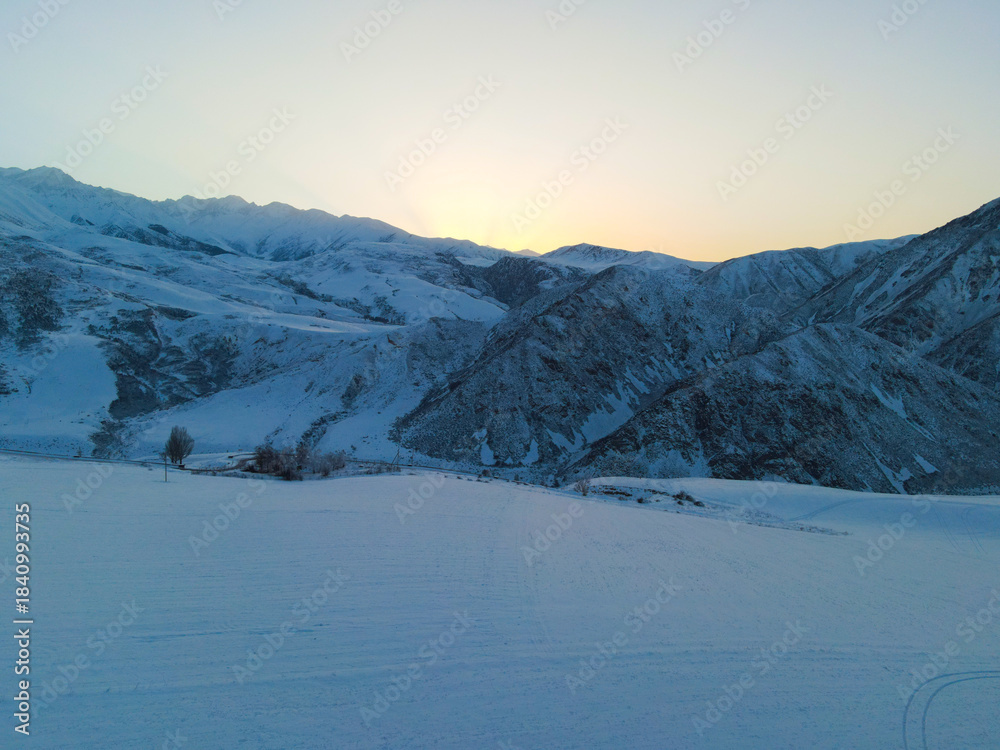 Fototapeta premium Wide aerial view of steep, snow-dusted mountains meeting a large, flat snow-covered field with a single tree and distant structures visible under a sunrise sky