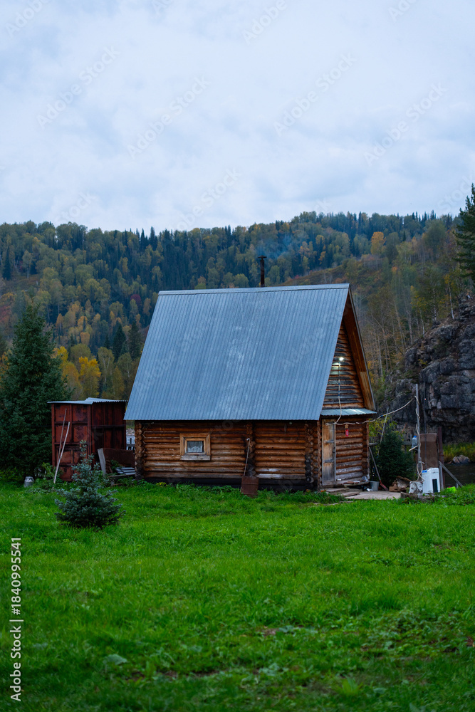Obraz premium A simple wooden cabin with a gray metal roof stands in a vibrant green meadow against a backdrop of dense, mixed forest covering a steep hillside under a cloudy sky