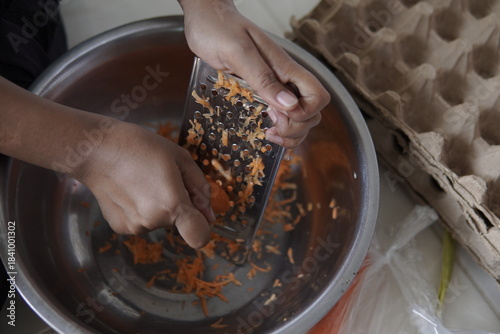 Hands grating fresh carrots into a metal bowl for cooking