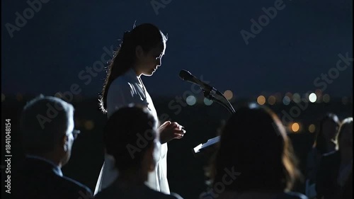 Woman delivering a speech at night with a microphone, surrounded by an audience against a city skyline