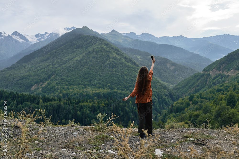 Naklejka premium A person stands on a rocky overlook, arms raised, gazing over distant mountains and lush landscape, capturing a moment of triumph in a vast outdoor scene.