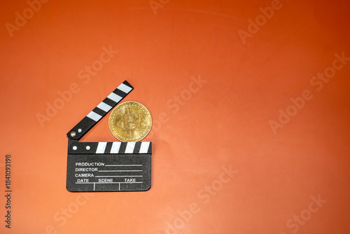 A Bitcoin coin rests on a film clapperboard against an orange background. This image suggests cryptocurrencies' potential in film production.