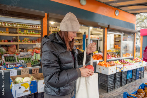 A woman is buying fresh vegetables and fruits at an outdoor market stall. She holds a pomegranate while placing her purchases into a reusable tote bag. The candid and natural scene reflects everyday s