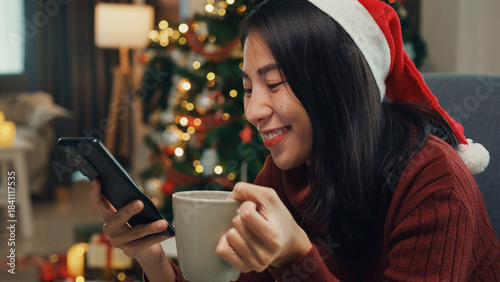 Positive Asia female wear red sweater new normal celebrate christmas smell aromatic coffee cup sit on wing chair check online greeting message from friend and family on phone in living room at house.