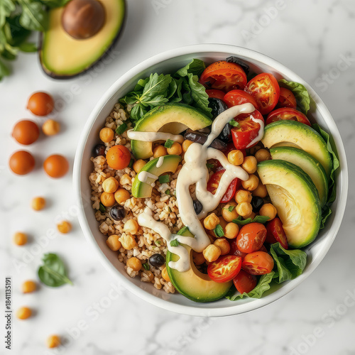 A white bowl holds a vibrant salad of greens, tomatoes, avocado, chickpeas, and black olives, accompanied by a halved avocado on a marble countertop.