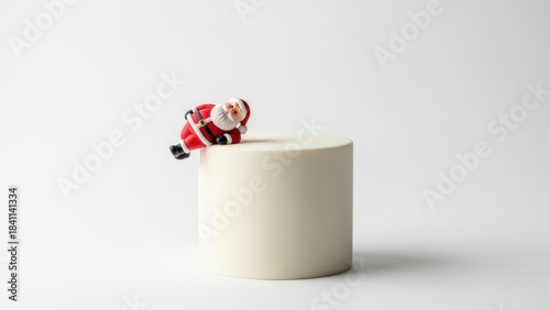 Minimalist Christmas cake with playful Santa figurine leaning over sleek white cylinder, modern festive dessert photography on clean background for elegant winter holiday celebrations


