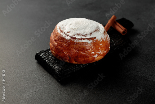 Round, delicious bun with cinnamon and powdered sugar on a black wooden board on a gray background