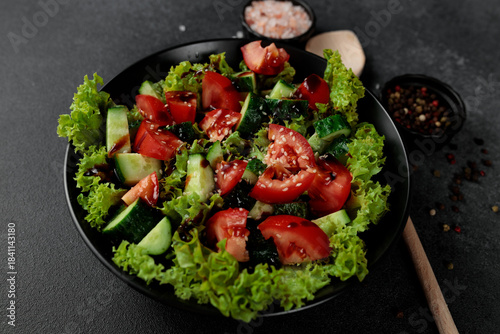 Vegetable salad of tomatoes, cucumbers, onions, lettuce dressed with aromatic dressing in a black plate on a gray background