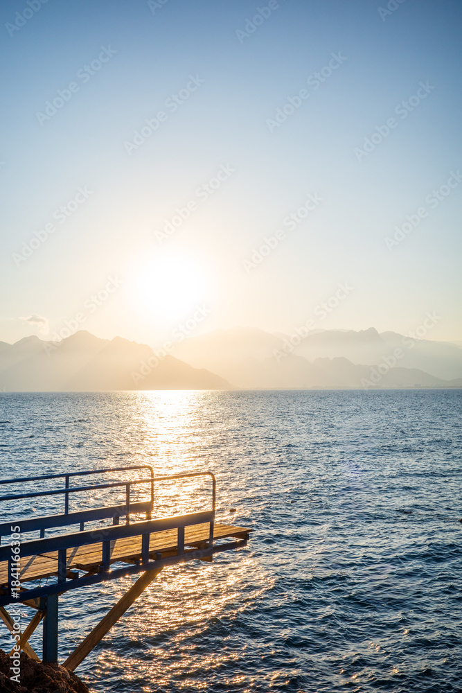 Obraz premium A peaceful sunset scene featuring a wooden pier overlooking calm ocean waters, with golden sunlight reflecting across the sea. Soft mountain silhouettes fade into the warm evening haze