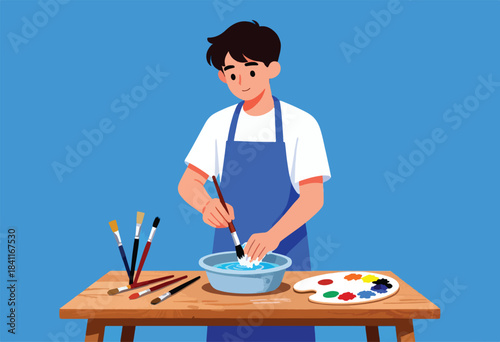 A person with a blue apron cleans a paintbrush over a bowl of water on a wooden table