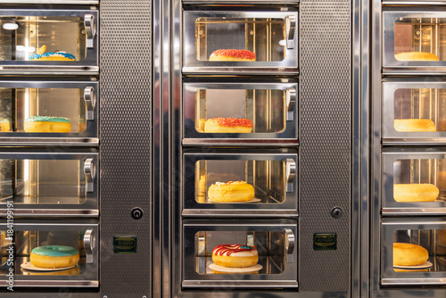 Automated vending machine filled with various colorful glazed donuts behind glass doors, offering sweet treats for sale in a public shopping area.