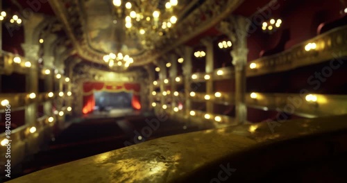 A view from the balcony of a grand theater featuring ornate architecture, golden chandeliers, and rows of red seating. Soft lighting creates a warm and inviting atmosphere for performances.