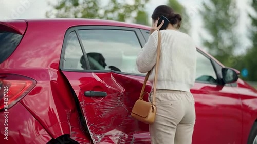 A woman is standing next to a red car that has significant damage to its side, appearing to be the result of a collision. She is on the phone, likely reporting the accident.