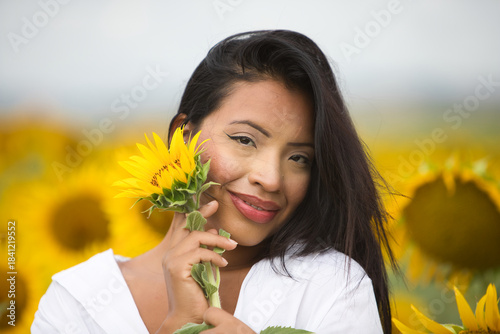 Fotografie A beautiful mulatto Latin woman with a white shirt brings it close to her face and flirts with a sunflower