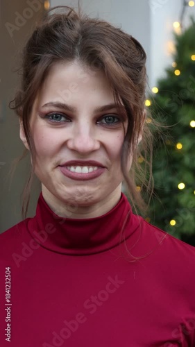 Close-up portrait of an attractive young woman smiling and laughing outdoors. She has an elegant updo hairstyle and wears a red turtleneck, standing near a lit Christmas tree.