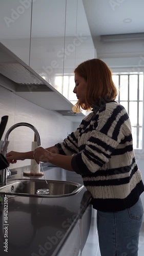 Red-haired woman doing household chores and washing a glass in the kitchen sink with running water
