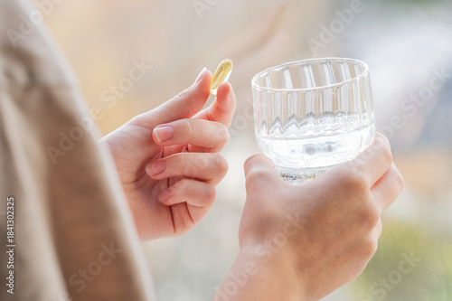 A close-up of a person's hands holding yellow softgel in one palm and a glass of water in the other.