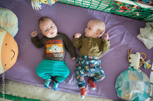 Two cute infants, twins brothers, lie side-by-side in a purple crib, wearing colorful outfits and socks. Stuffed animals and blankets surround them in their sleeping space
