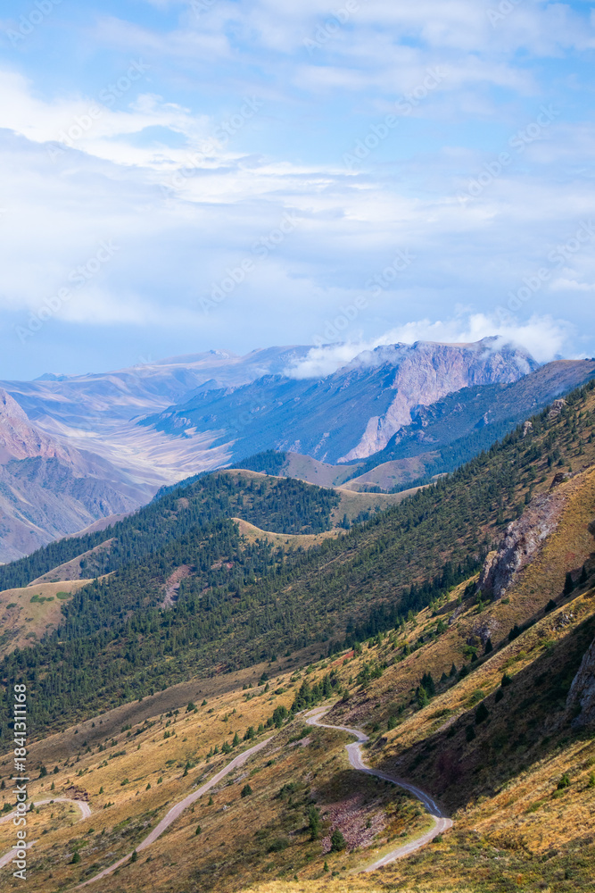 Fototapeta premium Picturesque landscape of a dirt road cutting across a steep green mountain slope with rugged peaks and clouds in the distance.