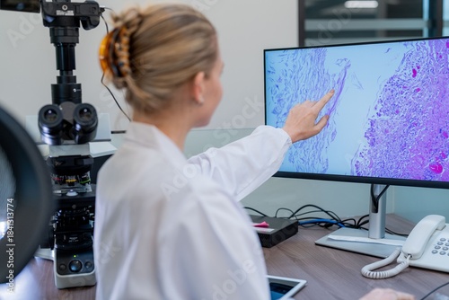 A scientist wearing a lab coat points at a computer screen showing a tissue sample. The laboratory is equipped with a microscope and other tools. The focus is on analysis and research.