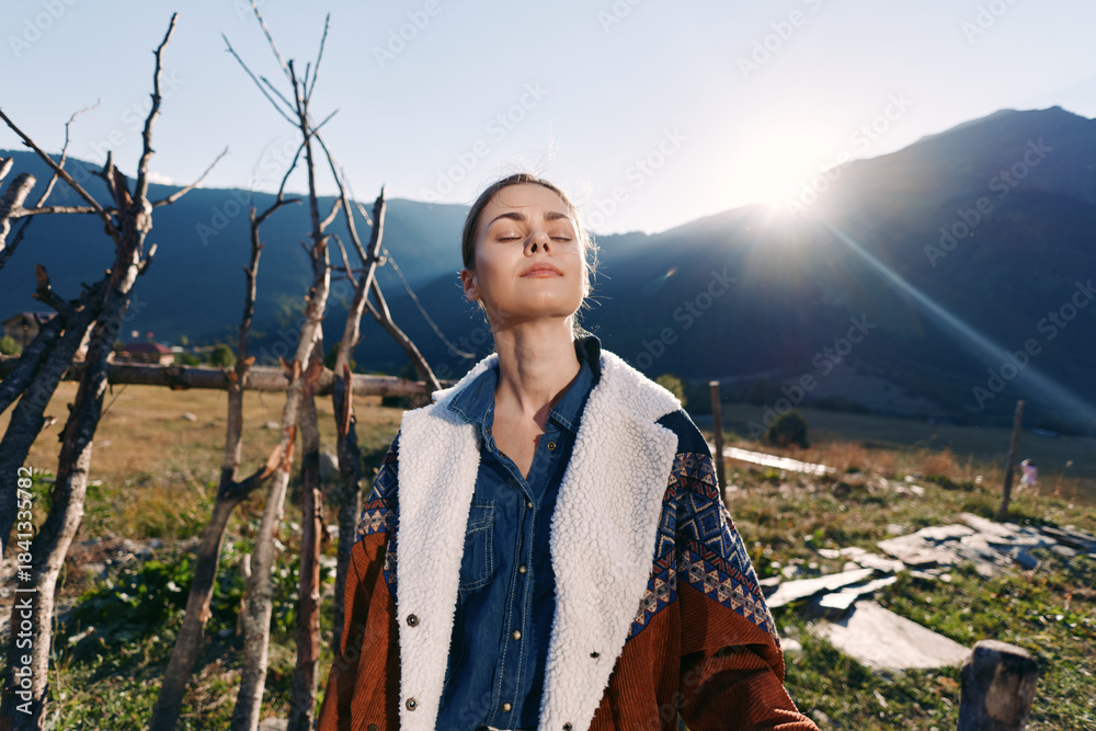 Naklejka premium Woman standing in mountains with sunlit outdoors scene, enjoying nature in a meadow wearing a warm jacket, eyes closed in relaxation and peaceful outdoor portrait at golden hour