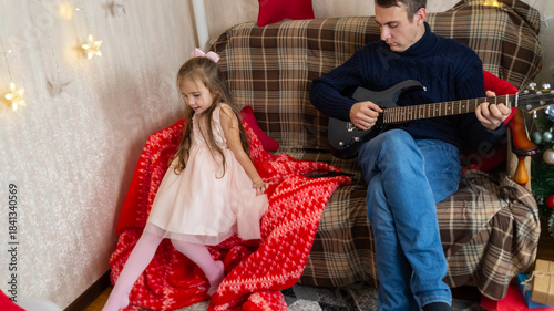 Father and little girl have fun together on Christmas holidays. Dad plays the guitar for his daughter.