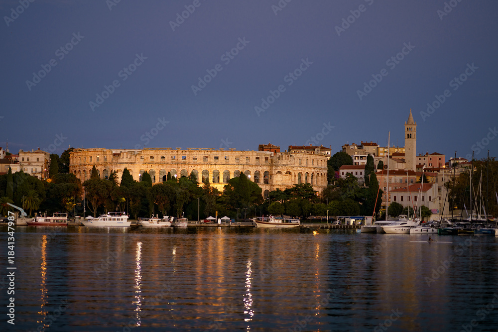 Fototapeta premium Evening view of Pula with the illuminated Roman Amphitheatre, calm marina, boats and reflections on the Adriatic Sea. Scenic coastal cityscape of Croatia.
