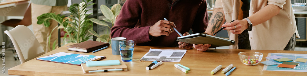 Obraz premium Caucasian young adult woman and Black young adult man collaborating at desk, reviewing documents and digital tablet, surrounded by office supplies and paperwork in modern workspace